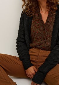 Kvinna som bär bruna högmidjade byxor, en brun mönstrad blus med röda detaljer och en svart stickad kofta, sittande med händerna vilande på benen.