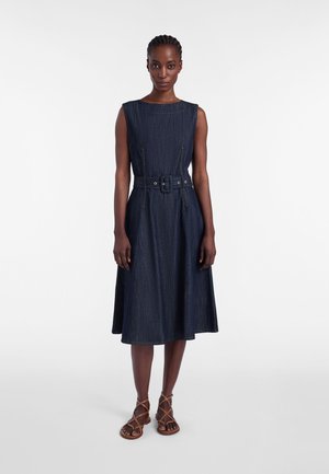 Nainen pukeutuneena hihattomaan tummansiniseen denimmekkoon, jossa on vyö, ja ruskeisiin remmikenkiin, seisomassa valkoista taustaa vasten.