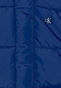 Tmavě modrá péřová bunda s prošitými částmi, na levé horní straně malým logem CK vyšitým bílou a modrou nití. Hladká textura.