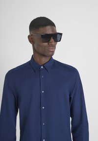 Occhiali da sole neri quadrati con finitura opaca, indossati insieme a una camicia blu marinara con bottoni. La camicia presenta un colletto strutturato e bottoni lungo la parte anteriore.