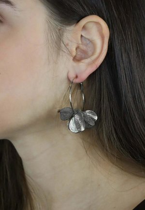 Srebrne kolczyki koła z warstwowymi, teksturowanymi kwiatowymi wzorami, z misternymi detalami na płatkach, prezentowane na uchu modelki.