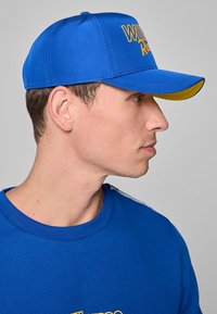 Niebieska czapka z żółtymi akcentami, z zakrzywionym daszkiem i perforowanym wierzchem dla wentylacji. Napis "Win Racing" w pogrubionej żółtej czcionce.