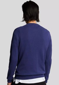 Maglione blu navy con scollatura tonda, maniche lunghe e polsini a coste. Texture liscia, design aderente, indossato sopra una camicia bianca.