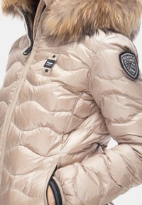 Giacca puffer beige con design trapuntato, cappuccio foderato in pelliccia, chiusura con zip e un patch ricamato sulla spalla. Finitura textured e lucida.