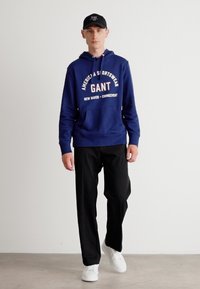 Sudadera azul con logo blanco de GANT, pantalones negros y zapatillas blancas. El modelo lleva una gorra negra a juego. Diseño sencillo, ajuste casual.
