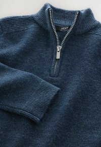 Granatowy sweter wełniany z prążkowanym wysokim kołnierzem i mankietami, metalowy zamek błyskawiczny na pół, a także widoczna metka marki wewnątrz kołnierza.