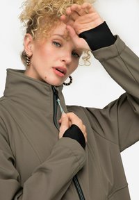 Giacca softshell verde oliva con colletto alto, zip anteriore, dettagli neri sui polsini e tessuto testurizzato. Presenta un cordino alla zip.