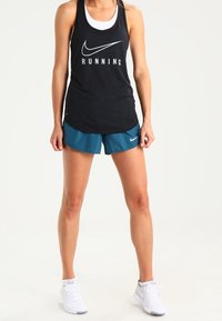 Femme portant un débardeur de course noir Nike, un short Nike turquoise et des chaussures de course blanches Nike, debout devant un fond clair.