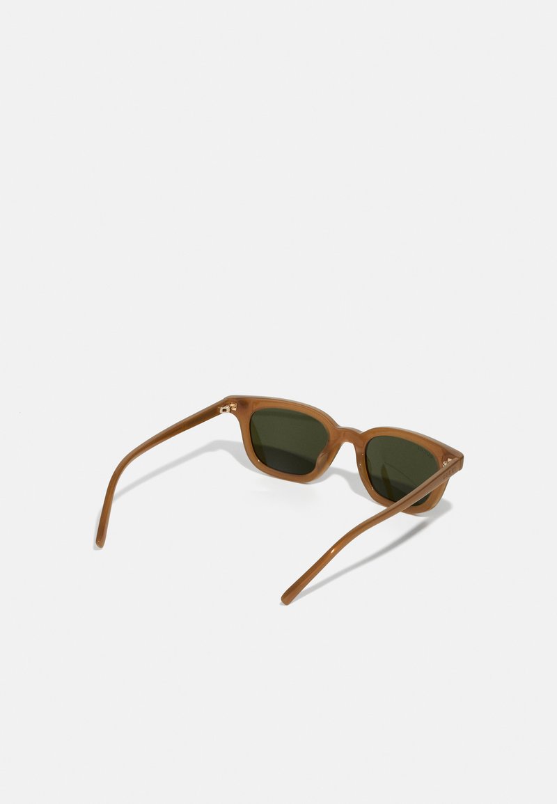 Occhiali da sole di plastica marrone con lenti verdi, caratterizzati da un design angolare ispirato al vintage e bracci sottili, posati ad un angolo su uno sfondo bianco.