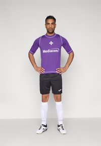 Fioletowa koszulka piłkarska z krótkimi rękawami, z białym logo. Czarne spodenki z białymi dodatkami oraz białe skarpety do kolan z wzorem.