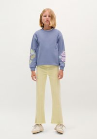 Felpa azzurra con motivi floreali sulle maniche, abbinata a pantaloni giallo chiaro a palazzo e scarpe bianche. Outfit semplice e casual.