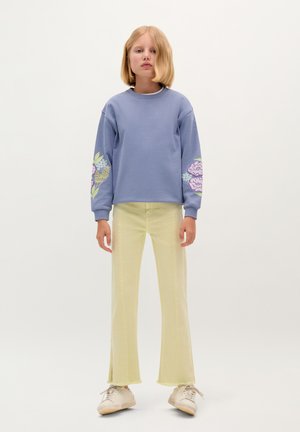 Světle modrá mikina se květinovými vzory na rukávech, v kombinaci se světle žlutými rozšířenými kalhotami a bílými botami. Jednoduchý, casual outfit.
