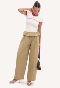 Femme portant un T-shirt blanc cintré avec une bordure rouge et un texte, pantalon beige à jambes larges, sandales blanches à talons, tenant un sac à main marron.