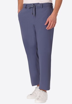 Uomo con pantaloni blu con coulisse e scarpe da ginnastica bianche, in piedi con una mano in tasca su sfondo uniforme.