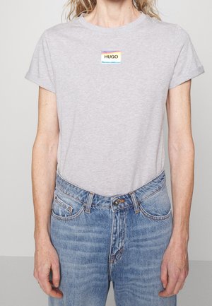 Person wearing light gray t-shirt with a rainbow "HUGO" logo patch and high-waisted blue jeans with rolled sleeves.