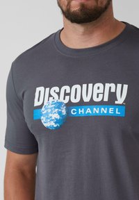 Grijze katoenen t-shirt met de tekst "Discovery CHANNEL" in wit en blauw, vergezeld van een blauwe aardbol graphic. Ronde hals en korte mouwen.