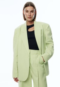 Lichtgroene oversized blazer van textuurstof, gecombineerd met een zwarte top met een uitgesneden detail. Losse bijpassende broek.