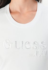Balta trumpa rankovėmis marškinėliai su išsiuvinėtu "Guess" logotipu, puoštas deimantais ir "est. 1981" kontrastinga balta tekstūra. Apvali iškirptė.