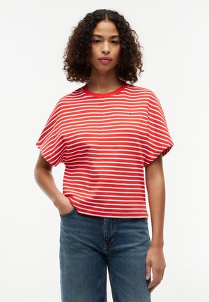Donna con capelli ricci che indossa una maglia a maniche corte a righe rosse e bianche e jeans blu, in piedi con una mano in tasca su sfondo bianco.