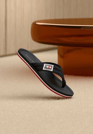 Μαύρο σαγιονάρι με κόκκινη και λευκή σόλα και υφασμάτινο λουράκι με ορθογώνιο λογότυπο, τοποθετημένο σε μπεζ χαλί κοντά σε καφέ τραπέζι.