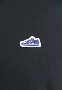 Parchment bordado de zapatilla en azul y blanco sobre un fondo de tela negra. El parche presenta detalles en los cordones y en el logo.