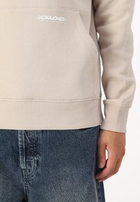 Felpa beige chiaro con tasca a marsupio e polsini a coste. Presenta il logo bianco "JACK & JONES" sul petto. Abbinata a pantaloni shorts in denim.