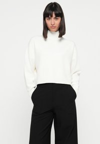 Weißer, verkürzter Rollkragenpullover mit geripptem Saum, kombiniert mit schwarzen, weit geschnittenen Hosen. Glatte Textur, lässige Passform, minimalistisches Design.