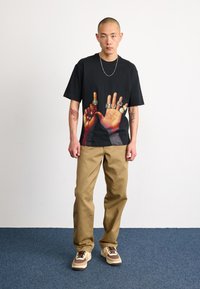 Černé tričko s grafikou rukou nosících prsteny, béžové kalhoty a vícebarevné tenisky na modrém koberci. Viditelný jednoduchý řetízek.