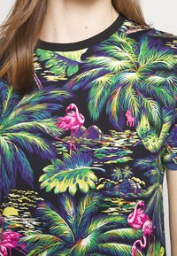 Černé tričko s krátkým rukávem s živým tropickým potiskem, zahrnujícím palmové listy, růžové plameňáky a barevné akcenty v zelené, modré a žluté.