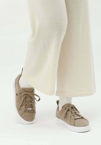 Beige mocka sneakers med tonade snören och en vit gummisula, ihop med krämfärgade vida byxor och vita ribbade strumpor.