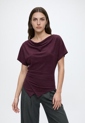 Jeune femme aux longs cheveux bruns portant un haut drapé bordeaux et un pantalon large gris foncé, debout devant un fond clair uni.