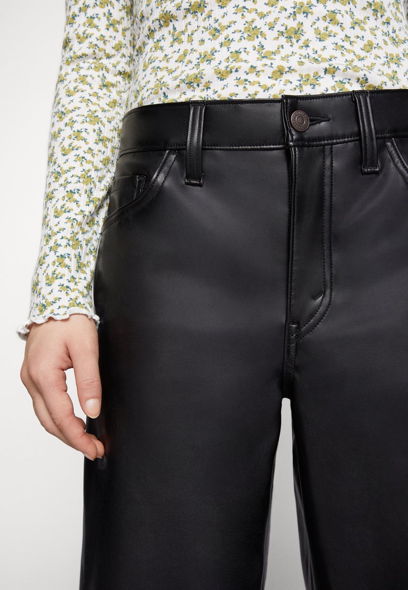 Persoon in zwarte hoge faux leren broek en een witte blouse met lange mouwen met een klein geel en groen bloemenpatroon.