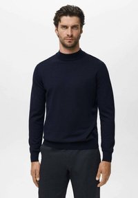 Man met krullend haar in een strakke donkerblauwe coltrui en zwarte broek, die tegen een effen lichte achtergrond staat.
