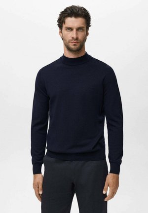 Man with curly hair wearing a fitted dark navy turtleneck sweater and black trousers, standing against a plain light background.