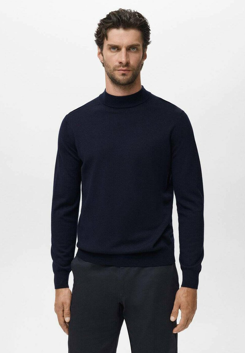 Hombre con cabello rizado que lleva un suéter de cuello alto ajustado de color azul marino oscuro y pantalones negros, de pie contra un fondo claro y liso.
