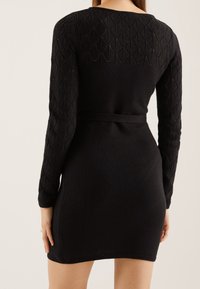 Model shows a fitted black dress with long, textured sleeves and a tied waist, highlighting a sleek silhouette against a neutral background.
