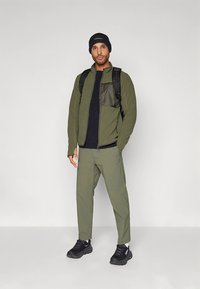 Flis jakna maslinasto zelene boje sa patentnim zatvaračem i džepom sprijeda, uparena s crnom majicom i maslinasto zelenim hlačama. Crne tenisice i kapa.