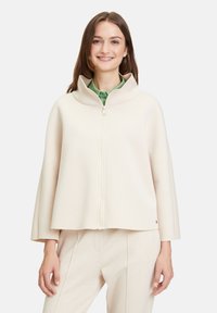 Giacca zip-up di colore crema con colletto alto, caratterizzata da una silhouette squadrata e una texture liscia. Design semplice con dettagli minimi.
