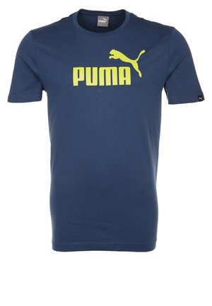 Homme portant un T-shirt bleu Puma, un short gris Puma et des baskets blanches, debout devant un fond blanc uni.