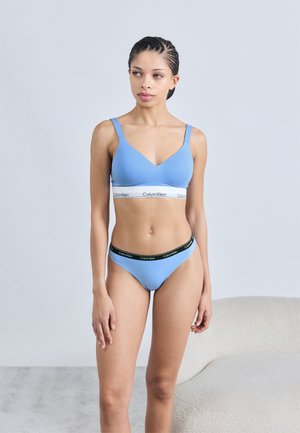 Lyseblåt sæt med bralette og g-streng med v-hals design. Har et hvidt Calvin Klein logo bånd. Glat tekstur, minimalistisk stil.