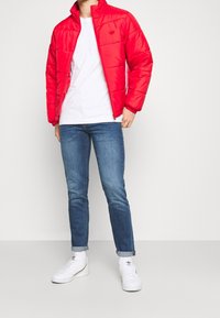 Red padded jacket with high collar, two side pockets, worn over a white t-shirt. Paired with blue jeans and white sneakers.