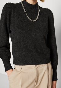 Kobieta ubrana w ciemnoszary, puszysty sweter z długim rękawem, srebrny naszyjnik łańcuszkowy oraz beżowe, wysokogatunkowe spodnie z rękami w kieszeniach.