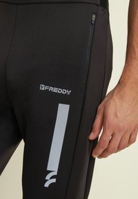 Pantalons de sport noirs avec une poche zippée, un logo gris et des accents géométriques. Fabriqués en tissu lisse et extensible avec un design ajusté.