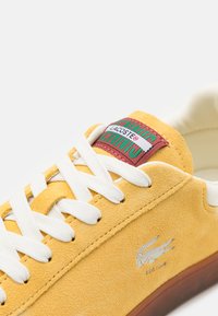 Žlté semišové tenisky Lacoste s bielymi šnúrkami, červenou a zelenou nášivkou na jazyku a strieborným logo krokodíla na boku.