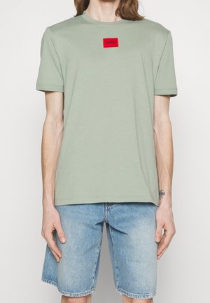 Helles grünes Baumwoll-T-Shirt mit Rundhalsausschnitt, kurzen Ärmeln und rotem rechteckigem Logo-Aufnäher. Getragen mit hellblauen Jeansshorts.