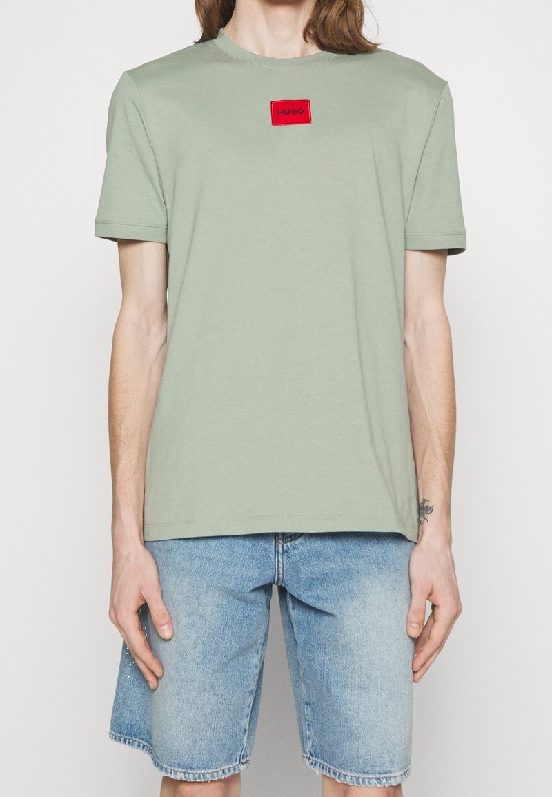 Camiseta de algodón verde claro con cuello redondo, mangas cortas y parche de logo rectangular rojo. Combinada con pantalones cortos de denim azul claro.