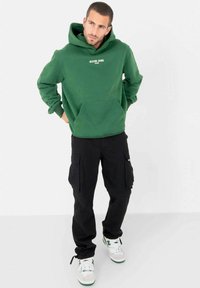 Groene hoodie met een voorzak en witte tekst, gecombineerd met zwarte cargobroeken. Model draagt witte sneakers met groene accenten.