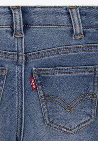 Jean en denim bleu avec couture visible, deux poches arrière et une petite étiquette rouge. La texture semble lisse avec un léger dégradé.