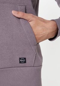 Lila Kapuzenpullover aus Baumwollmischung, mit einer Fronttasche und einem kleinen schwarzen Bench-Label auf der unteren linken Seite. Insgesamt glatte Textur.