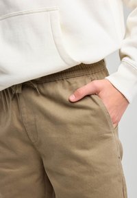 Cremefarbene Kapuzenjacke mit einer Vorderstasche, kombiniert mit beigen, elastischen Hosen mit seitlichen Taschen und einer strukturierten Stoffoberfläche.